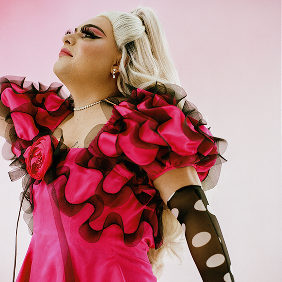 Aniel Gonzalez in a pink gown with black trimmed ruffles and long, black gloves with holes in them, stands looking up and to the left, in front of a pink ombre background. 
