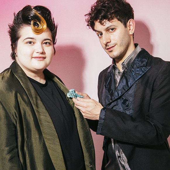 For pride prom, Alex Tudela on the right, pins a blue fabric floral corsage to Lukas Petroski's khaki green jacket lapel. Lukas also wears a black t-shirt.