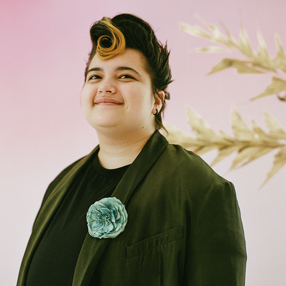 For pride prom, Lukas Petroski wears an olive green suit jacket with a turquoise blue green fabric flower brooch on his lapel with a black t-shirt underneath. There is a pink ombre background and some beige floral branches behind him. 
