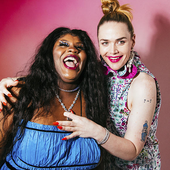 For pride prom, Willie Norris wears pearl and diamond chandelier earrings, a sleeveless, ruffle-neck printed and magenta dress, an emerald and diamond ring, and diamond bracelets, on the right. She has her arms around Taylor Collins Allen, on the left. Taylor wears a blue, ruched gown with black trim. and a diamond necklace that Willie is touching. 