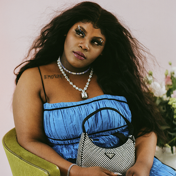 Taylor Collins Allen sits in a green velvet chair with a pink ombre background. Taylor wears a blue, ruched gown with black trim, a diamond necklace with a silver-tone lock pendant, and is holding a crystal-covered Prada nylon shoulder bag.